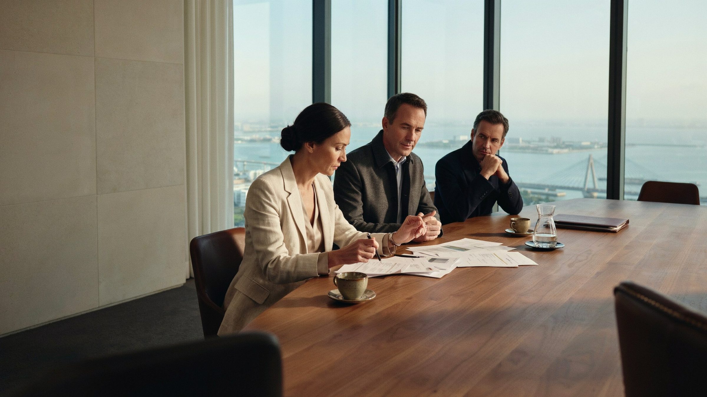 Three senior executives reviewing documents at a walnut boardroom table overlooking the water and a cable-stayed bridge